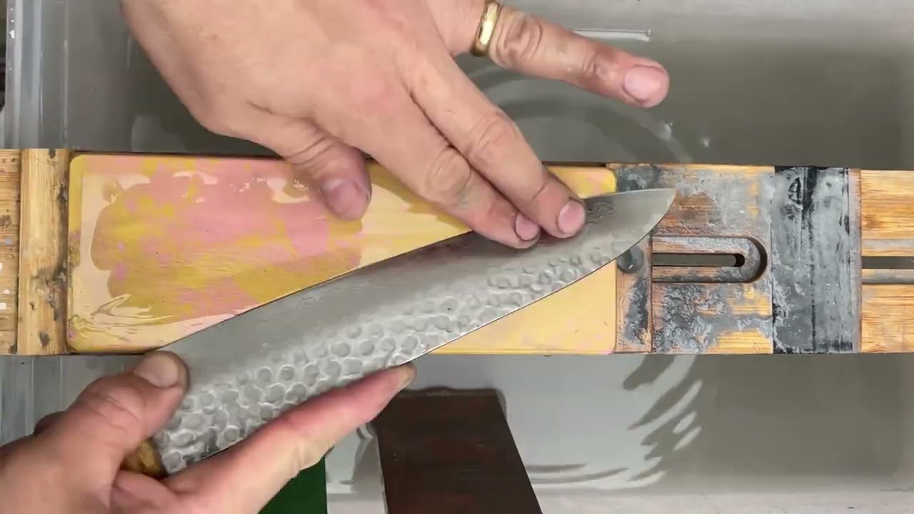 A hammered blade drawn across a wet whetstone, fingertips controlling the pressure